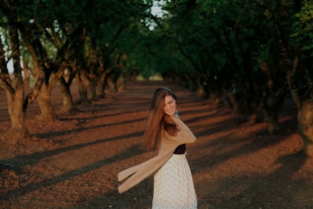 A woman in a long skirt and sweater is twirling joyfully on a path lined with trees on either side. The setting is bathed in warm, golden sunlight, and the shadows of the trees create a pattern on the ground. The atmosphere is peaceful and serene, surrounded by abundant greenery.