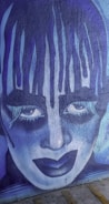 A striking mural featuring a stylized face with exaggerated makeup, painted in shades of blue and purple. The artistic expression includes intense eyes and dramatic makeup, giving it a theatrical and somewhat haunting appearance.