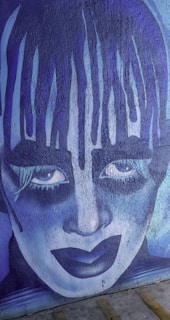 A striking mural featuring a stylized face with exaggerated makeup, painted in shades of blue and purple. The artistic expression includes intense eyes and dramatic makeup, giving it a theatrical and somewhat haunting appearance.
