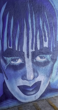 A striking mural featuring a stylized face with exaggerated makeup, painted in shades of blue and purple. The artistic expression includes intense eyes and dramatic makeup, giving it a theatrical and somewhat haunting appearance.