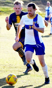 Two soccer players are in action on the field, intensely focused on the game. The player in the white and blue uniform is leading and appears to be in control of the ball. The player behind, in a dark blue and yellow uniform, is closely pursuing. Both players are captured in motion with expressions of determination.