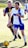 Two soccer players are in action on the field, intensely focused on the game. The player in the white and blue uniform is leading and appears to be in control of the ball. The player behind, in a dark blue and yellow uniform, is closely pursuing. Both players are captured in motion with expressions of determination.
