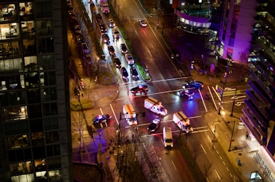 An aerial view of a city intersection at night shows several emergency vehicles with flashing lights blocking the crossroad. The streets are lined with parked cars, and the buildings on either side are illuminated, with one displaying several windows with lights on.
