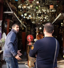 Two people are standing in front of a shop filled with an assortment of vintage lamps and chandeliers. The shop is cluttered with various antique items, including a red fan and ornate furniture. One person appears to be gesturing expressively while the other listens with crossed arms.