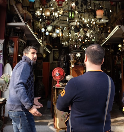 Two people are standing in front of a shop filled with an assortment of vintage lamps and chandeliers. The shop is cluttered with various antique items, including a red fan and ornate furniture. One person appears to be gesturing expressively while the other listens with crossed arms.