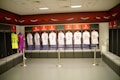 A locker room featuring a row of football jerseys hanging in open lockers. Each jersey is neatly displayed, with names and numbers visible. The room has a modern design with benches in front of the lockers. A mannequin is dressed in a goalkeeper uniform on the left, and a glass case on the right holds a football. The color theme includes red, white, and black accents.