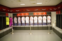 A locker room featuring a row of football jerseys hanging in open lockers. Each jersey is neatly displayed, with names and numbers visible. The room has a modern design with benches in front of the lockers. A mannequin is dressed in a goalkeeper uniform on the left, and a glass case on the right holds a football. The color theme includes red, white, and black accents.