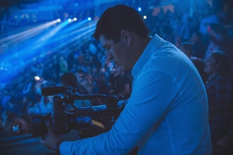 A person in a blue shirt operates a video camera in a dimly lit auditorium filled with a crowd. Blue lighting and beams add a dramatic atmosphere to the scene, and the focus is on capturing an event.