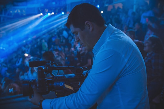 A person in a blue shirt operates a video camera in a dimly lit auditorium filled with a crowd. Blue lighting and beams add a dramatic atmosphere to the scene, and the focus is on capturing an event.