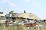 A small white and red airplane is parked in front of a building with a prominent aviation-related emblem. Three flags, including one with a green and yellow pattern, are flying near the building. The scene includes some grass in the foreground and a clear sky above.