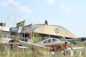 A small white and red airplane is parked in front of a building with a prominent aviation-related emblem. Three flags, including one with a green and yellow pattern, are flying near the building. The scene includes some grass in the foreground and a clear sky above.
