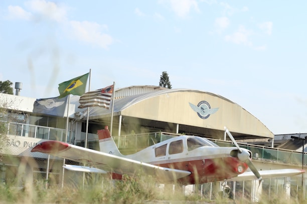 A small white and red airplane is parked in front of a building with a prominent aviation-related emblem. Three flags, including one with a green and yellow pattern, are flying near the building. The scene includes some grass in the foreground and a clear sky above.