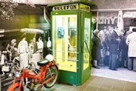 A green and yellow telephone booth with the word 'TELEFON' displayed at the top. Inside, there is a blue rotary payphone. In front of the booth is a red vintage motorcycle. The background consists of a black and white mural depicting a crowd of people, some standing and others engaging in various activities.