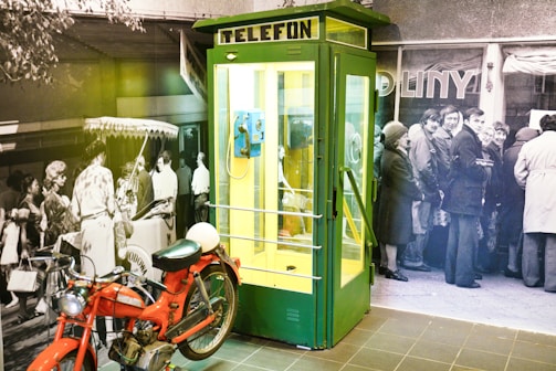 A green and yellow telephone booth with the word 'TELEFON' displayed at the top. Inside, there is a blue rotary payphone. In front of the booth is a red vintage motorcycle. The background consists of a black and white mural depicting a crowd of people, some standing and others engaging in various activities.