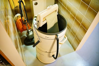 A vintage washing machine with a wringer is placed in a utility room. The machine is cylindrical with a wringer attachment on top, next to a tiled wall. Cleaning equipment like a mop and bucket are also visible in the corner.