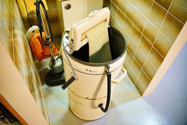 A vintage washing machine with a wringer is placed in a utility room. The machine is cylindrical with a wringer attachment on top, next to a tiled wall. Cleaning equipment like a mop and bucket are also visible in the corner.