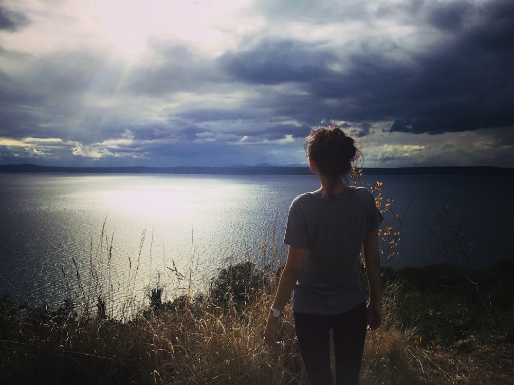 woman in gray t-shirt standing on green grass field near body of water during daytime