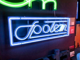 A neon sign displaying the word 'Społem' in bright blue neon lights against a dark background. Above it, there is a partial view of another sign with green neon, shaped like a letter or symbol. The setting appears to be an indoor space, possibly an exhibition or a storefront.