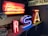 An indoor display of vintage neon signs, including letters in bright colors. The signs include glowing letters such as 'R', 'S', and 'A' in red, blue, and yellow respectively. Above, a larger red-lit sign spells out 'Perfumery'. The setting is dimly lit, enhancing the vibrancy of the neon lights.