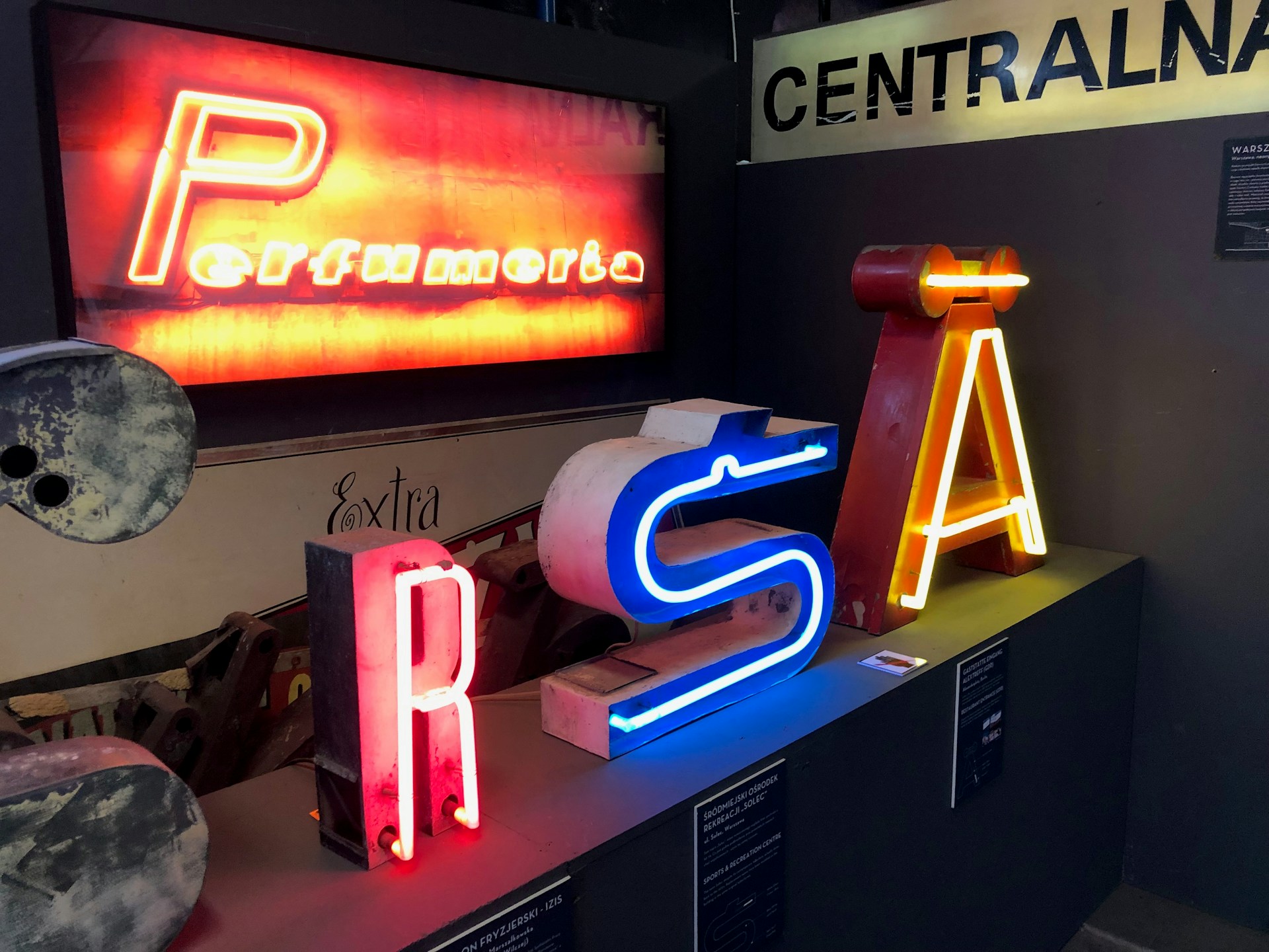 An indoor display of vintage neon signs, including letters in bright colors. The signs include glowing letters such as 'R', 'S', and 'A' in red, blue, and yellow respectively. Above, a larger red-lit sign spells out 'Perfumery'. The setting is dimly lit, enhancing the vibrancy of the neon lights.