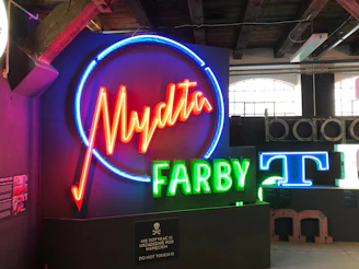 A collection of neon signs is displayed, featuring vibrant colors. The largest sign displays the word 'Mydła' in red neon surrounded by a blue circle. Below it, the word 'FARBY' is illuminated in green neon. Nearby, parts of other signs and letters are visible, including a blue 'T'. The signs are mounted against a backdrop of a dimly lit room with wooden beams and industrial-style windows.
