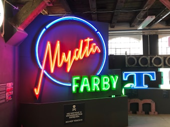 A collection of neon signs is displayed, featuring vibrant colors. The largest sign displays the word 'Mydła' in red neon surrounded by a blue circle. Below it, the word 'FARBY' is illuminated in green neon. Nearby, parts of other signs and letters are visible, including a blue 'T'. The signs are mounted against a backdrop of a dimly lit room with wooden beams and industrial-style windows.