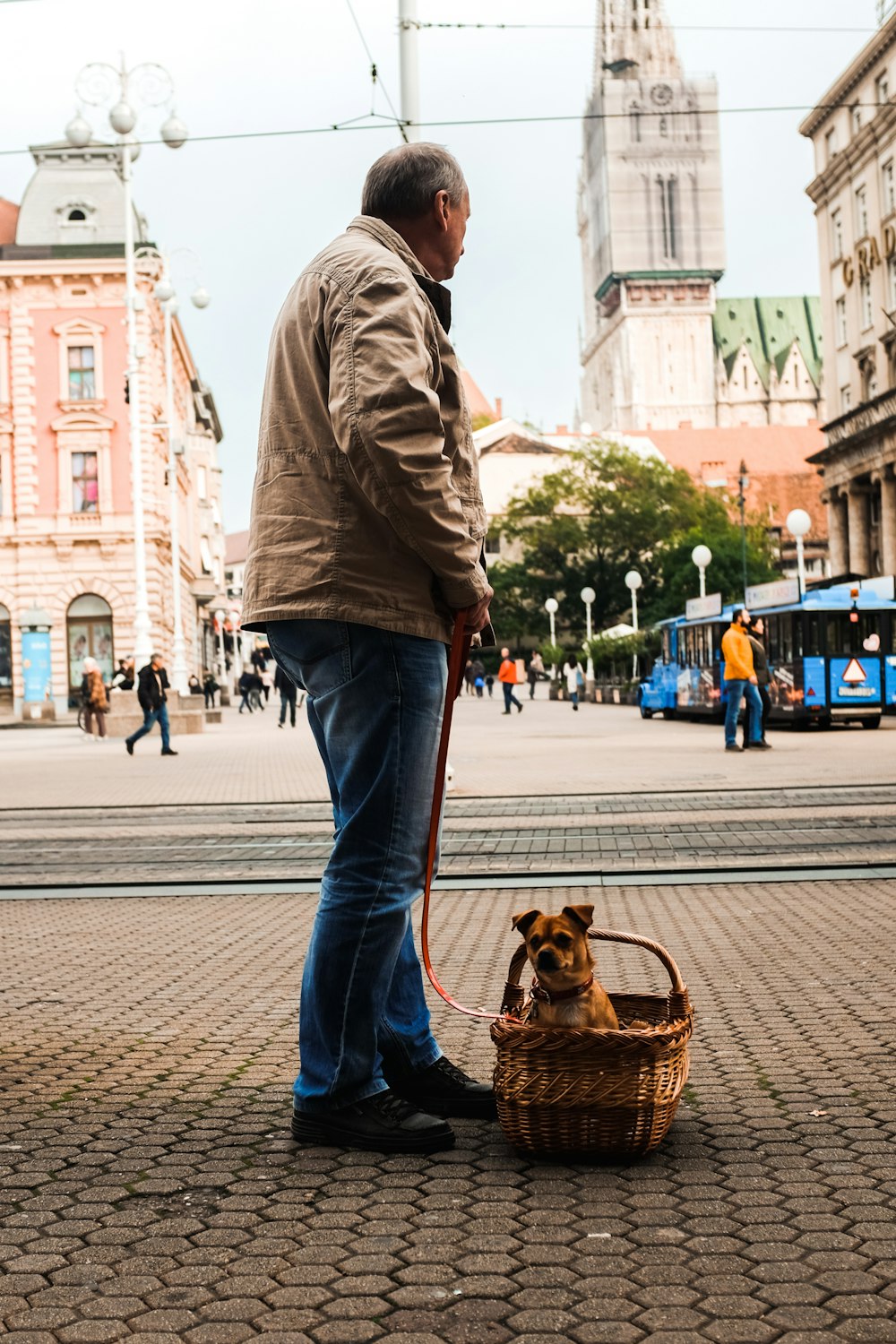 man in brown jacket and blue denim jeans holding brown dog on street during daytime