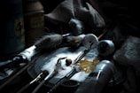 Art supplies are scattered on a surface, including various brushes, tubes of paint, jars, and a palette with paint smudges. The lighting is dim, highlighting the textures and details of the brushes and paint tubes, suggesting a workspace for painting.