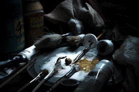 Art supplies are scattered on a surface, including various brushes, tubes of paint, jars, and a palette with paint smudges. The lighting is dim, highlighting the textures and details of the brushes and paint tubes, suggesting a workspace for painting.
