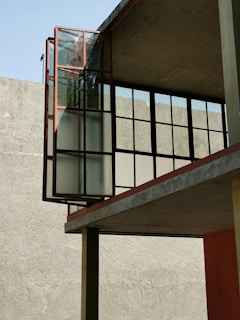 A modern architectural structure featuring large glass panels framed with metal that extend outward from a concrete building. The background consists of a plain grey wall and a clear blue sky.