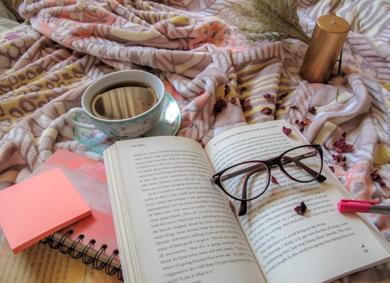 A cozy scene featuring an open book with a pair of glasses resting on it. A cup of coffee sits in a delicate floral-patterned cup and saucer nearby, alongside a notebook with a pink sticky note on top. A gold candle and dried flowers are arranged on a patterned blanket, creating a warm and inviting atmosphere.