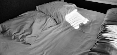 A neatly made bed with two pillows, covered in a plain, white fabric. Sunlight casts a rectangle of light across the bed from a nearby window, creating strong contrast and shadows.