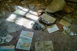 Scattered maps, papers, and a vintage suitcase lie on a dusty floor amidst debris and sunlight filtering through. The scene suggests abandonment and neglect, with worn and damaged items strewn about.