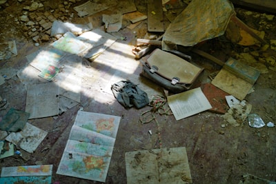 Scattered maps, papers, and a vintage suitcase lie on a dusty floor amidst debris and sunlight filtering through. The scene suggests abandonment and neglect, with worn and damaged items strewn about.