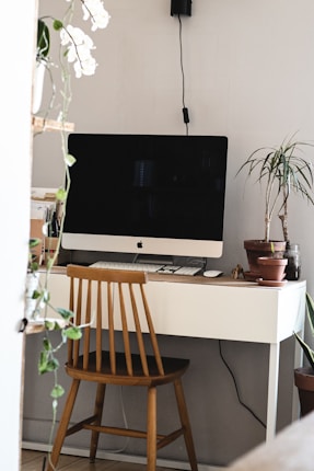 A minimalist workspace includes a sleek computer with a black screen on a white desk. A wooden chair is positioned in front of the desk. Potted plants and a few office supplies are situated around the setup, adding a decorative and functional touch.