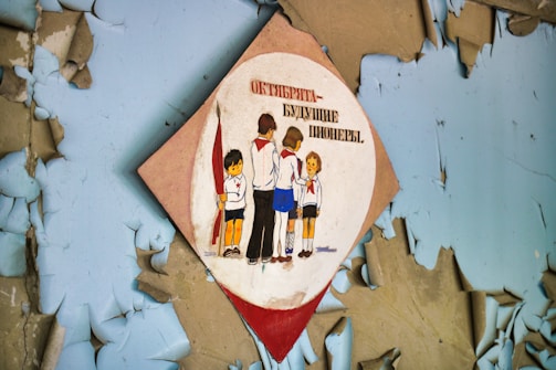 A deteriorated wall with peeling blue paint features a diamond-shaped sign. The sign depicts children in uniforms standing in a group, with one child holding a red flag. The text on the sign is in Cyrillic.