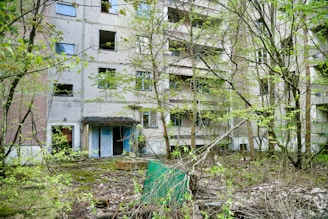 An abandoned multi-story building with broken windows and overgrown vegetation. The facade shows signs of decay and neglect, surrounded by trees and shrubs. The ground is covered with fallen leaves and debris, adding to the desolate and forgotten atmosphere.
