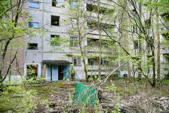 An abandoned multi-story building with broken windows and overgrown vegetation. The facade shows signs of decay and neglect, surrounded by trees and shrubs. The ground is covered with fallen leaves and debris, adding to the desolate and forgotten atmosphere.