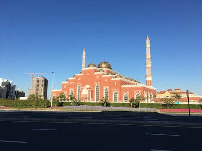 A large mosque features vibrant pink and white walls with intricate architectural details. Two tall minarets rise majestically against a clear blue sky. The surrounding area is landscaped with neatly maintained greenery and colorful flowerbeds. In the background, there are modern buildings and a construction crane, indicating development in the area.