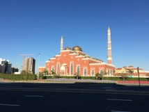 A large mosque features vibrant pink and white walls with intricate architectural details. Two tall minarets rise majestically against a clear blue sky. The surrounding area is landscaped with neatly maintained greenery and colorful flowerbeds. In the background, there are modern buildings and a construction crane, indicating development in the area.