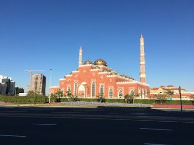 A large mosque features vibrant pink and white walls with intricate architectural details. Two tall minarets rise majestically against a clear blue sky. The surrounding area is landscaped with neatly maintained greenery and colorful flowerbeds. In the background, there are modern buildings and a construction crane, indicating development in the area.