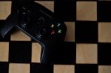 A black gaming controller with colorful buttons labeled X, Y, A, and B is placed on a wooden chessboard.