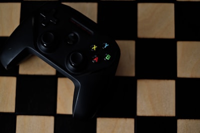 A black gaming controller with colorful buttons labeled X, Y, A, and B is placed on a wooden chessboard.