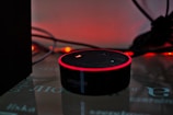 A smart speaker device with a circular shape sits on a reflective surface, highlighted by a red glowing ring on its top edge. Surrounding the speaker are tangled wires and a dark background with warm ambient lighting that gives a cozy yet minimalistic technological atmosphere.
