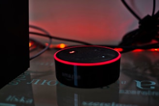 A smart speaker device with a circular shape sits on a reflective surface, highlighted by a red glowing ring on its top edge. Surrounding the speaker are tangled wires and a dark background with warm ambient lighting that gives a cozy yet minimalistic technological atmosphere.