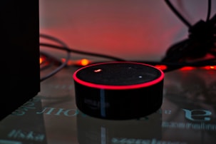 A smart speaker device with a circular shape sits on a reflective surface, highlighted by a red glowing ring on its top edge. Surrounding the speaker are tangled wires and a dark background with warm ambient lighting that gives a cozy yet minimalistic technological atmosphere.