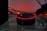 A smart speaker device with a circular shape sits on a reflective surface, highlighted by a red glowing ring on its top edge. Surrounding the speaker are tangled wires and a dark background with warm ambient lighting that gives a cozy yet minimalistic technological atmosphere.