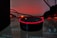A smart speaker device with a circular shape sits on a reflective surface, highlighted by a red glowing ring on its top edge. Surrounding the speaker are tangled wires and a dark background with warm ambient lighting that gives a cozy yet minimalistic technological atmosphere.
