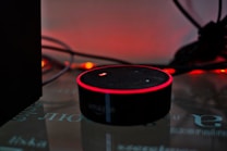 A smart speaker device with a circular shape sits on a reflective surface, highlighted by a red glowing ring on its top edge. Surrounding the speaker are tangled wires and a dark background with warm ambient lighting that gives a cozy yet minimalistic technological atmosphere.