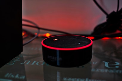 A smart speaker device with a circular shape sits on a reflective surface, highlighted by a red glowing ring on its top edge. Surrounding the speaker are tangled wires and a dark background with warm ambient lighting that gives a cozy yet minimalistic technological atmosphere.